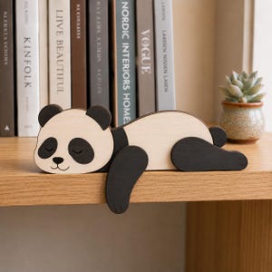 Puede incluir: Una figura de panda de madera plana con un cuerpo de color beige claro y orejas, parches oculares, extremidades y una nariz pequeña de color marrón oscuro. El panda está en posición de dormir con las patas delanteras sobre un borde.