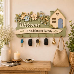 Pode incluir: Um suporte de parede para chaves em madeira com base verde sálvia, um ícone de casa com porta azul e detalhes florais. A placa exibe o texto Welcome e The Johnson Family. Inclui cinco ganchos dourados com várias chaves e um pingente dourado.