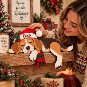 Könnte beinhalten: Ein Holzausschnitt eines schlafenden Beagle-Hundes, der eine rote Weihnachtsmütze mit dem in rotem Faden gestickten Namen Coco und einen grün-rot gestreiften Schal trägt. Der Hund hat braune, schwarze und weiße Fellzeichnungen.