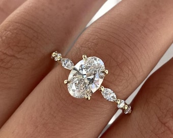 May include: A gold ring featuring an oval-cut diamond as the centerpiece. The band is adorned with smaller, round and marquise-cut diamonds. The ring is set against a background of a person's skin.