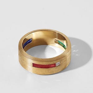 May include: A gold-colored ring with a brushed finish. The ring features a red rectangular stone, a small square diamond, and blue and green rectangular stones. The ring is set against a white background.