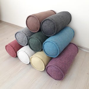 May include: A selection of cylindrical bolster cushions in diverse colours, such as red, white, yellow, green, blue, and purple. The cushions are stacked, displaying their rounded ends and textured fabric. The cushions are approximately 30 cm long.