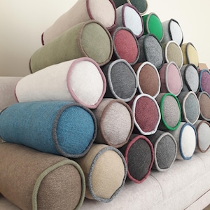 May include: A collection of cylindrical bolster pillows in various colors, including blue, green, beige, and gray. Each pillow has a contrasting trim and is arranged in a stacked display. The pillows are made of a textured fabric.