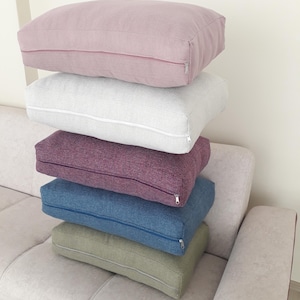 May include: Five rectangular cushions in a stack, in shades of dusty rose, light grey, deep purple, denim blue, and olive green. Each has a zip closure. They are stacked on a beige sofa.
