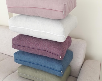 May include: A stack of five rectangular floor cushions featuring a textured fabric finish. The cushions are coloured in shades of dusty pink, light grey, deep purple, denim blue, and sage green, each equipped with a matching side zipper closure.