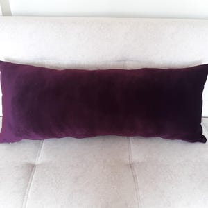 Könnte beinhalten: Ein langes, rechteckiges Kissen aus violettem Samt auf einem weißen Sofa.