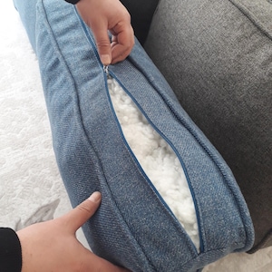 May include: A blue textured fabric cushion cover with a visible zip closure. The cover is partially unzipped, revealing a white, fluffy, fibrous filling material inside. The fabric has a woven appearance with a consistent blue colour throughout.