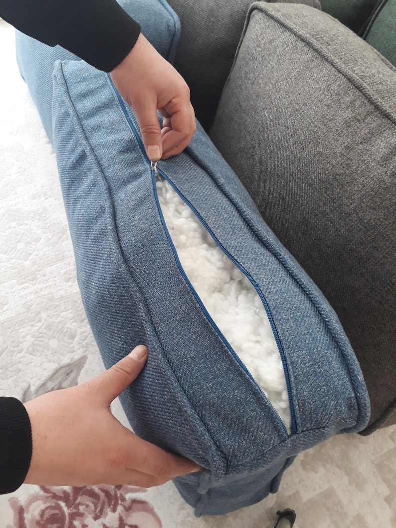 May include: A blue textured fabric cushion cover with a visible zip closure. The cover is partially unzipped, revealing a white, fluffy, fibrous filling material inside. The fabric has a woven appearance with a consistent blue colour throughout.