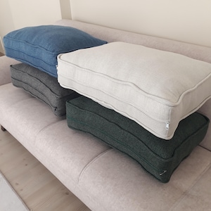 May include: Four rectangular floor cushions with a textured fabric finish. The set includes one blue, one light grey, one cream, and one dark green cushion. Each cushion features a piped edge detail and a visible side zip closure for the cover.