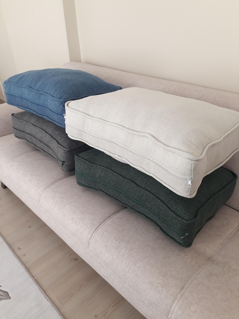 May include: Four rectangular floor cushions with a textured fabric finish. The set includes one blue, one light grey, one cream, and one dark green cushion. Each cushion features a piped edge detail and a visible side zip closure for the cover.