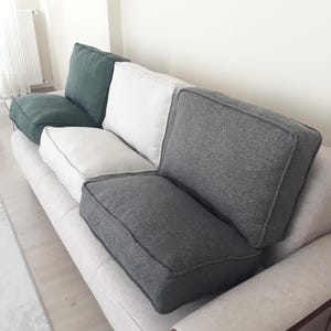 May include: A sofa featuring a light beige base with a set of removable cushions in dark green, white, and charcoal grey. Each cushion has a structured rectangular shape with visible piping along the edges, providing a layered seating arrangement.