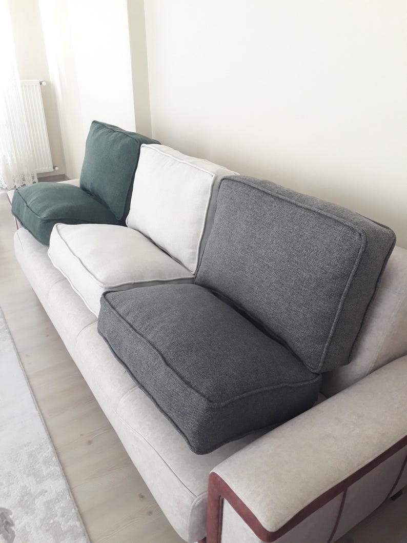 May include: A sofa featuring a light beige base with a set of removable cushions in dark green, white, and charcoal grey. Each cushion has a structured rectangular shape with visible piping along the edges, providing a layered seating arrangement.
