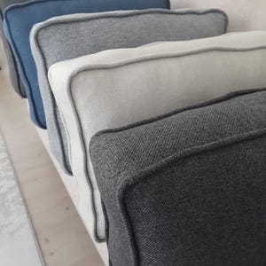 May include: A row of five square throw cushions arranged in a line, featuring a textured fabric finish with piped edges. The colours include dark charcoal, light cream, medium grey, muted blue, and a deep black, all displayed in a sequence.