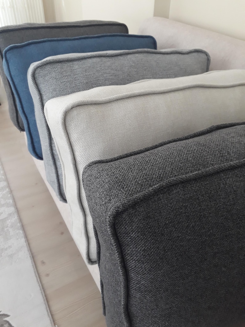 May include: A row of five square throw cushions arranged in a line, featuring a textured fabric finish with piped edges. The colours include dark charcoal, light cream, medium grey, muted blue, and a deep black, all displayed in a sequence.