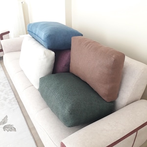 May include: A collection of rectangular throw pillows in various solid colors including blue, white, purple, dark green, and brown, stacked and arranged on a light beige sofa with a subtle textured fabric and thin pink piping along the armrest.
