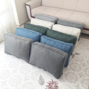 Può includere: Una collezione di cuscini rettangolari in vari colori, tra cui grigio, blu, verde e bianco. I cuscini sono disposti davanti a un divano beige, suggerendo un tema di arredamento per la casa. I cuscini hanno un design semplice e moderno.