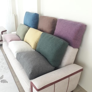 May include: A light-coloured sofa featuring a series of rectangular throw pillows in various solid colours including blue, brown, purple, olive green, yellow, white, dark green, grey, and light pink, arranged along the backrest and seat.