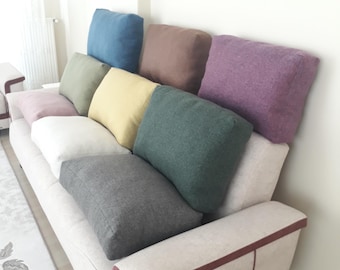 May include: A light-coloured sofa featuring a series of rectangular throw pillows in various solid colours including blue, brown, purple, olive green, yellow, white, dark green, grey, and light pink, arranged along the backrest and seat.