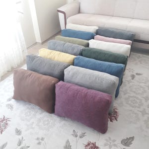 Könnte beinhalten: Eine Sammlung rechteckiger Kissen in verschiedenen Farben, darunter Braun, Lila, Grau, Blau und Gelb. Die Kissen sind auf einem floral gemusterten Teppich vor einem beigen Sofa angeordnet. Die Kissen scheinen aus einem strukturierten Stoff zu sein.