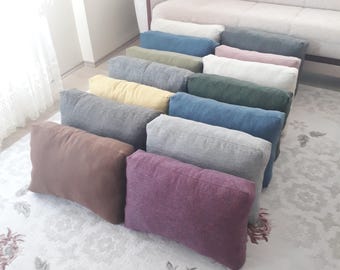 May include: A collection of rectangular throw cushions in various solid colours including brown, purple, grey, yellow, blue, green, pink, and off-white. The cushions feature a textured fabric finish and are arranged in rows on a patterned floor.
