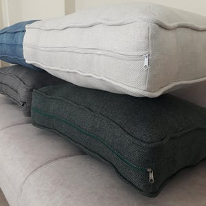May include: A stack of four rectangular floor cushions featuring textured fabric covers. The top cushion is off-white, followed by a blue cushion, a light grey cushion, and a dark charcoal grey cushion at the bottom, each with a visible side zipper.