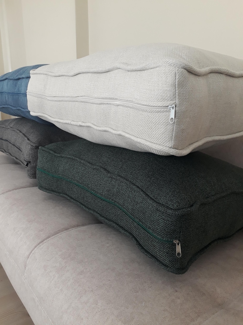 May include: A stack of four rectangular floor cushions featuring textured fabric covers. The top cushion is off-white, followed by a blue cushion, a light grey cushion, and a dark charcoal grey cushion at the bottom, each with a visible side zipper.