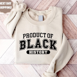 Op de afbeelding: Een beige sweatshirt met de tekst "PRODUCT OF BLACK HISTORY" in zwarte blokletters. De sweatshirt is op een houten oppervlak gevouwen. De mouwen zijn opgerold.