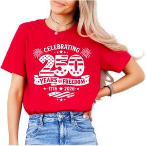 May include: A bright red short-sleeved t-shirt featuring a white graphic print that reads CELEBRATING 250 YEARS OF FREEDOM 1776-2026. The design includes large numbers filled with stars and stripes, firework icons, and a waving American flag motif.