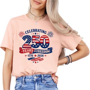 May include: A peach-colored t-shirt featuring a graphic with the text CELEBRATING 250 YEARS OF FREEDOM 1776-2026. The design includes the number 250 in a red, white, and blue American flag pattern, surrounded by fireworks and star accents.