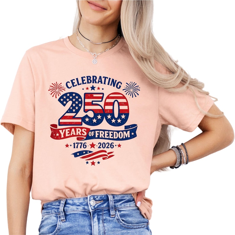 May include: A peach-colored t-shirt featuring a graphic with the text CELEBRATING 250 YEARS OF FREEDOM 1776-2026. The design includes the number 250 in a red, white, and blue American flag pattern, surrounded by fireworks and star accents.