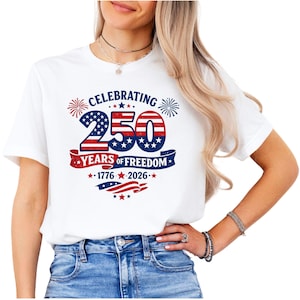 May include: A white short-sleeved t-shirt featuring a graphic with the text CELEBRATING 250 YEARS OF FREEDOM 1776-2026. The design includes red, white, and blue colors, star patterns, fireworks, and a waving flag motif centered on the chest area.