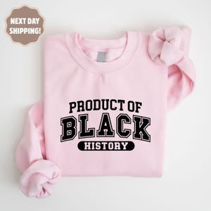 Op de afbeelding: Een lichtroze sweatshirt met de tekst "PRODUCT OF BLACK HISTORY" in een vetgedrukte, zwarte, collegiale lettertype. De sweatshirt heeft een ronde hals en lange mouwen.