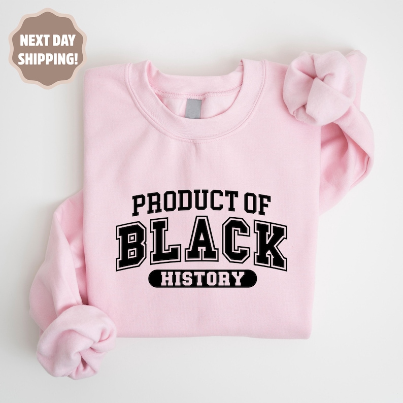 Op de afbeelding: Een lichtroze sweatshirt met de tekst "PRODUCT OF BLACK HISTORY" in een vetgedrukte, zwarte, collegiale lettertype. De sweatshirt heeft een ronde hals en lange mouwen.