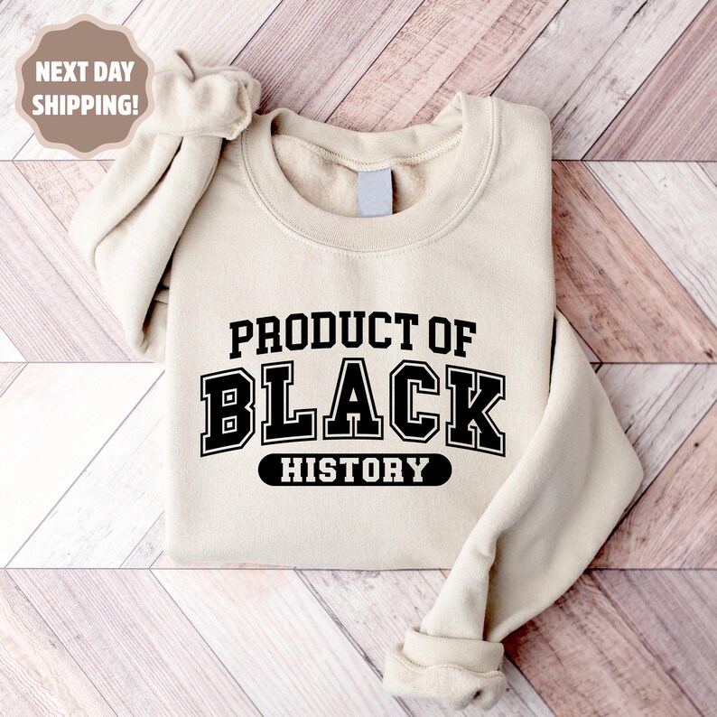 Op de afbeelding: Een beige sweatshirt met de tekst "PRODUCT OF BLACK HISTORY" in zwarte blokletters. De sweatshirt is op een houten oppervlak gevouwen. De mouwen zijn opgerold.