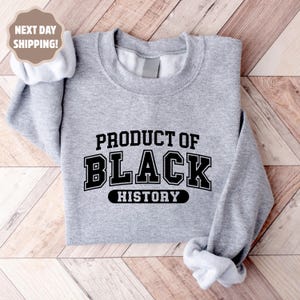 Op de afbeelding: Een lichtgrijs sweatshirt met de tekst "PRODUCT OF BLACK HISTORY" in zwarte blokletters. De sweatshirt heeft een ronde hals en lange mouwen. De afbeelding bevat ook een "NEXT DAY SHIPPING!" afbeelding.