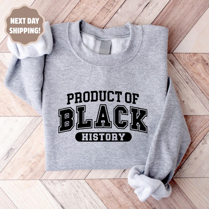 Op de afbeelding: Een lichtgrijs sweatshirt met de tekst "PRODUCT OF BLACK HISTORY" in zwarte blokletters. De sweatshirt heeft een ronde hals en lange mouwen. De afbeelding bevat ook een "NEXT DAY SHIPPING!" afbeelding.