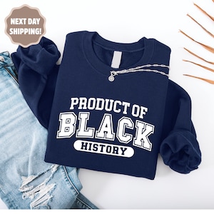 Op de afbeelding: Een marineblauw sweatshirt met de witte tekst "PRODUCT OF BLACK HISTORY". Een zilveren ketting met een hanger is over het sweatshirt gedrapeerd. Het sweatshirt wordt gepresenteerd op een wit oppervlak.