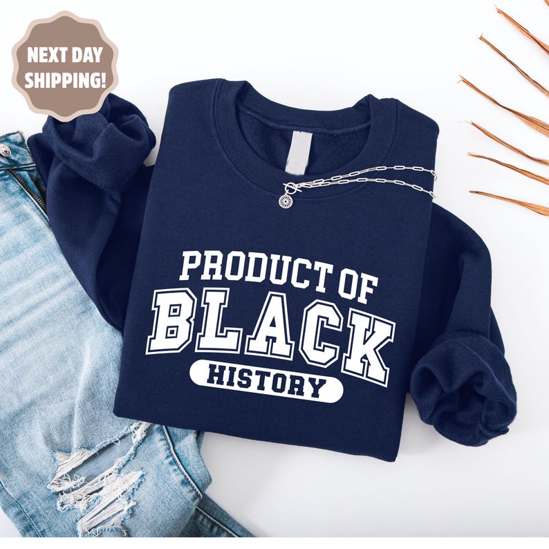 Op de afbeelding: Een marineblauw sweatshirt met de witte tekst "PRODUCT OF BLACK HISTORY". Een zilveren ketting met een hanger is over het sweatshirt gedrapeerd. Het sweatshirt wordt gepresenteerd op een wit oppervlak.