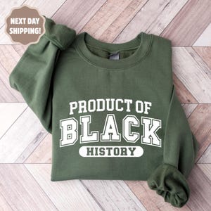 Op de afbeelding: Een olijfgroene sweatshirt met de witte tekst "PRODUCT OF BLACK HISTORY". De sweatshirt is opgevouwen op een lichtbruin houten oppervlak. De mouwen zijn opgerold. De tekst is vetgedrukt.