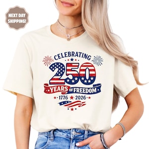 May include: A cream-colored short-sleeve t-shirt featuring a graphic design with the text CELEBRATING 250 YEARS OF FREEDOM 1776-2026. The design includes red, white, and blue patriotic elements, stars, stripes, and fireworks illustrations.