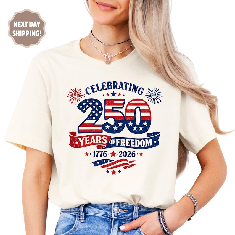 May include: A cream-colored short-sleeve t-shirt featuring a graphic design with the text CELEBRATING 250 YEARS OF FREEDOM 1776-2026. The design includes red, white, and blue patriotic elements, stars, stripes, and fireworks illustrations.