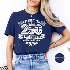 May include: A navy blue short-sleeve t-shirt featuring a white graphic print that reads CELEBRATING 250 YEARS OF FREEDOM 1776-2026. The design includes a large number 250 with stars and stripes patterns, firework icons, and a waving flag banner.