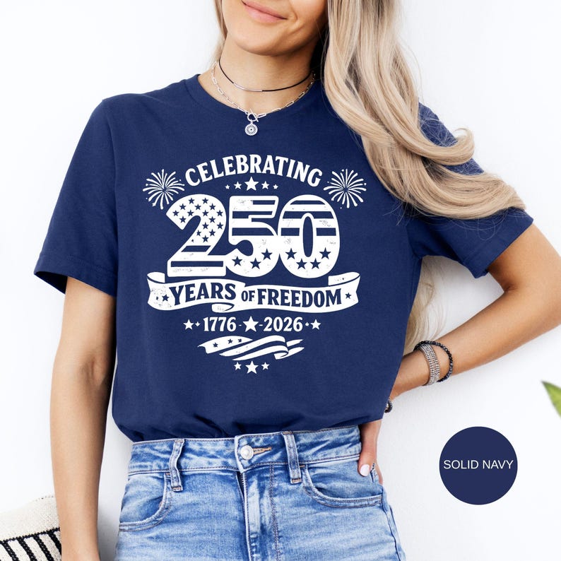May include: A navy blue short-sleeve t-shirt featuring a white graphic print that reads CELEBRATING 250 YEARS OF FREEDOM 1776-2026. The design includes a large number 250 with stars and stripes patterns, firework icons, and a waving flag banner.