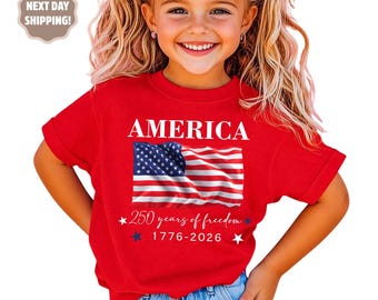 May include: A red t-shirt featuring the word "AMERICA" above a graphic of the American flag. Below the flag, the text reads "250 years of freedom 1776-2026" with star accents. The shirt is worn by a child.