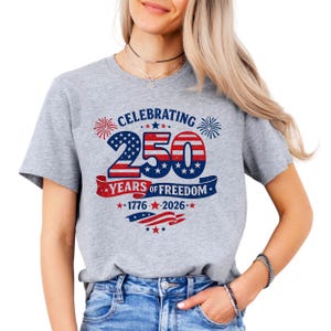 May include: A heather grey t-shirt featuring a graphic design with the text CELEBRATING 250 YEARS OF FREEDOM 1776-2026. The design includes red, white, and blue colors, star patterns, fireworks, and a waving flag motif in a distressed, vintage style.