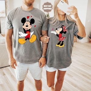 A man and a woman are standing together, both wearing t-shirts with Mickey Mouse designs. The man is holding the woman's hand, and they are both smiling.