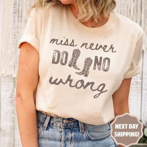 May include: Cream-colored t-shirt with the phrase "miss never do no wrong" in a distressed, brown font. The design includes two cowboy boot illustrations. The shirt is paired with blue jeans.