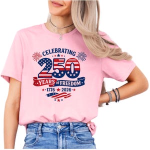 May include: A light pink short-sleeved t-shirt featuring a graphic design with the text CELEBRATING 250 YEARS OF FREEDOM 1776-2026. The graphic includes the number 250 in a red, white, and blue American flag pattern, fireworks, and stars.