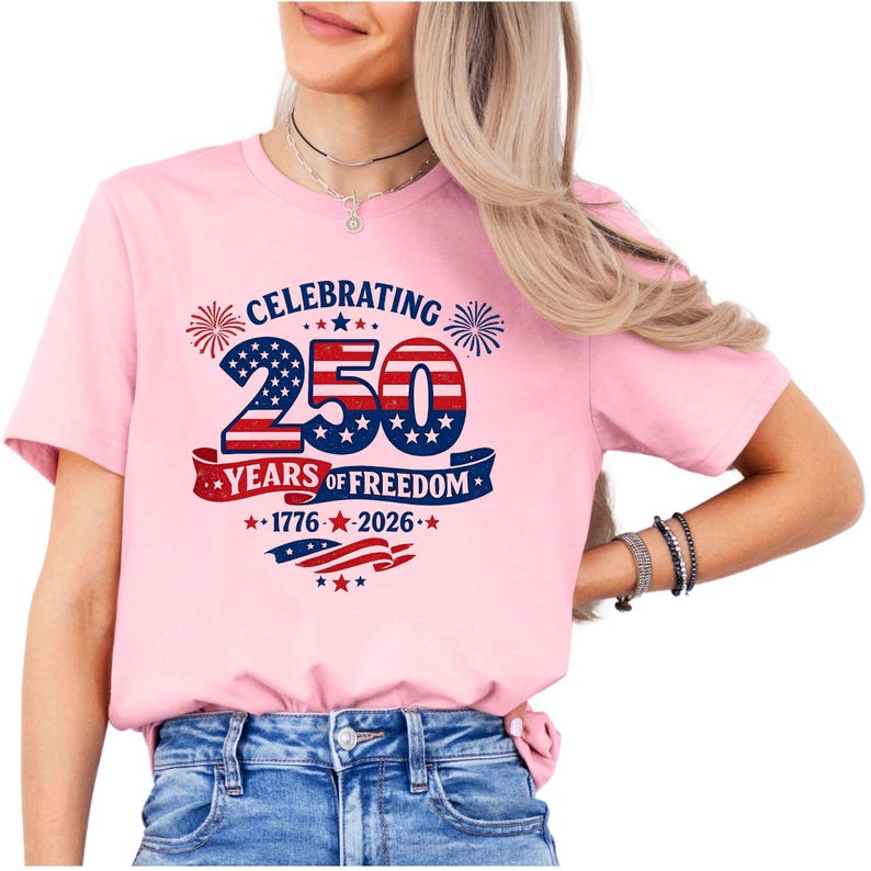 May include: A light pink short-sleeved t-shirt featuring a graphic design with the text CELEBRATING 250 YEARS OF FREEDOM 1776-2026. The graphic includes the number 250 in a red, white, and blue American flag pattern, fireworks, and stars.