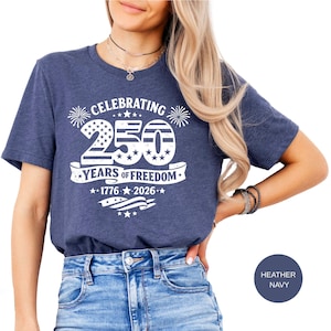 May include: A heather navy blue short-sleeve t-shirt featuring white graphic text that reads CELEBRATING 250 YEARS OF FREEDOM 1776 2026. The design includes a large number 250 with stars and stripes patterns, firework icons, and a waving flag.